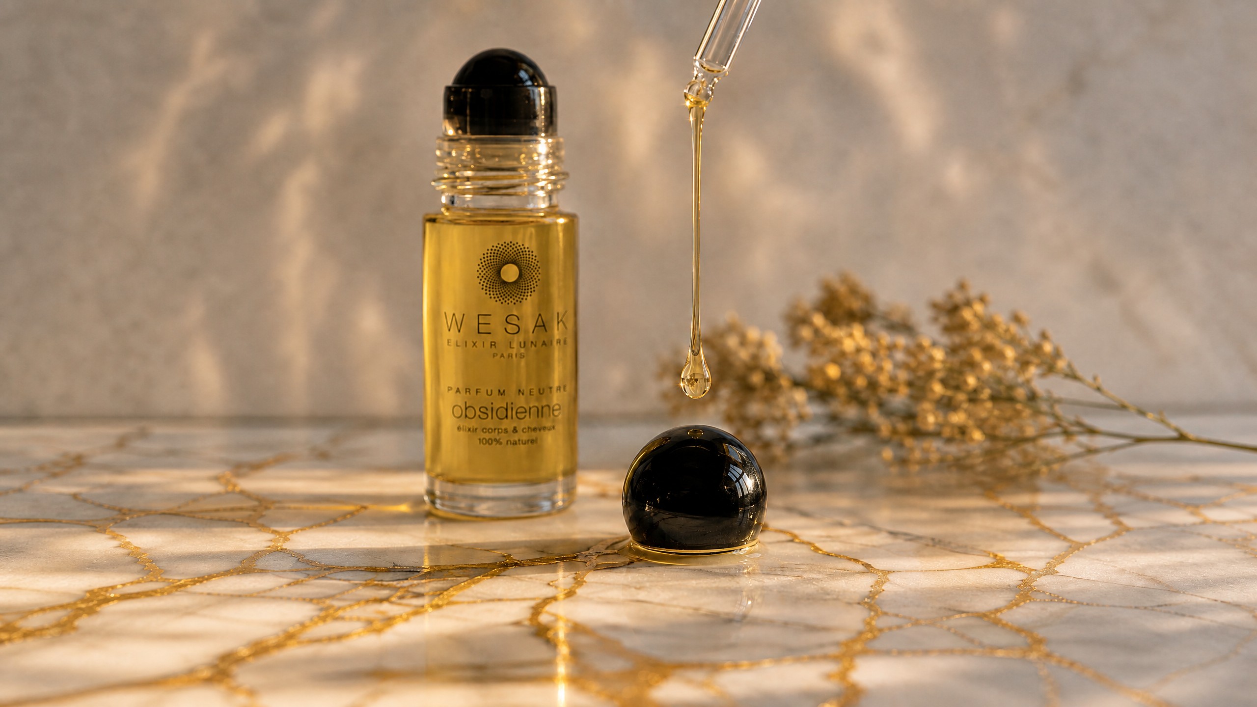 Élixir obsidienne Wesak Paris Élixir Lunaire Paris sur marbre blanc veiné d'or avec bille obsidienne noire et goutte d'huile dorée - roll-on cristal soin corps et cheveux